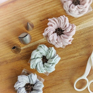 以下が含まれることがあります： 木製の表面に、バタークリームの花の装飾が4つあります。花はピンク、パープル、グリーンの色合いです。表面には、絞り出し口金が3つと白いハサミが1組あります。