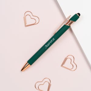 K&ouml;nnte beinhalten: Ein dunkelgr&uuml;ner Stift mit goldenen Akzenten und dem Namen "Aurora" auf der Seite. Der Stift liegt auf einer hellrosa Oberfl&auml;che, zusammen mit drei herzf&ouml;rmigen B&uuml;roklammern.