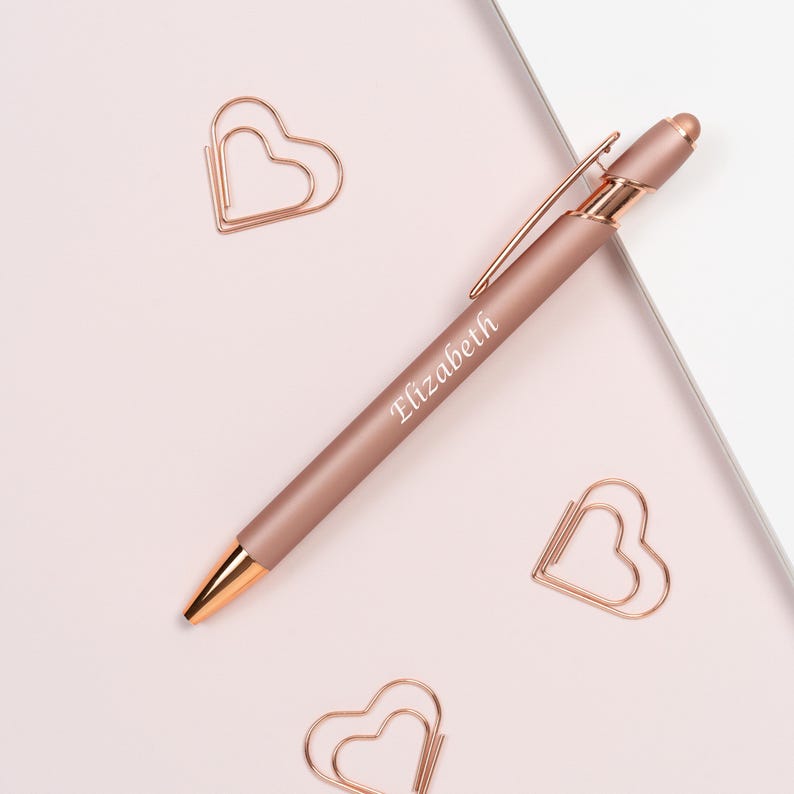 K&ouml;nnte beinhalten: Ein ros&eacute;goldener Stift mit dem eingravierten Namen "Elizabeth" und drei herzf&ouml;rmigen B&uuml;roklammern. Der Stift hat eine goldfarbene Spitze und einen Clip, vor einem hellrosa Hintergrund.