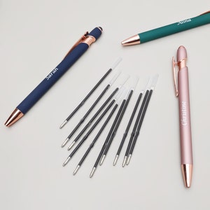 May include: Three personalised pens with rose gold accents. The pens are blue, green, and pink. The names "Sarah", "Anna", and "Christine" are engraved on the pens. The image also shows a pile of black and clear pen refills.