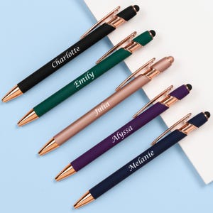 K&ouml;nnte beinhalten: Eine Auswahl an personalisierten Stiften in Schwarz, Gr&uuml;n, Ros&eacute;gold, Lila und Marineblau. Jeder Stift hat eine goldfarbene Spitze und einen Clip, mit einem auf dem Schaft aufgedruckten Namen. Die Stifte sind diagonal auf einer hellblauen Oberfl&auml;che angeordnet.