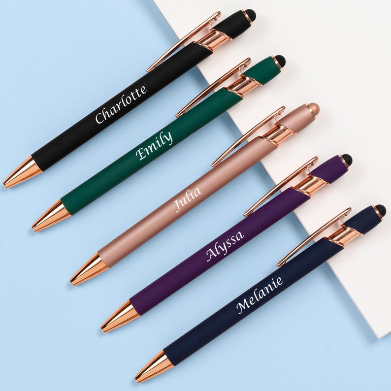 K&ouml;nnte beinhalten: Eine Auswahl an personalisierten Stiften in Schwarz, Gr&uuml;n, Ros&eacute;gold, Lila und Marineblau. Jeder Stift hat eine goldfarbene Spitze und einen Clip, mit einem auf dem Schaft aufgedruckten Namen. Die Stifte sind diagonal auf einer hellblauen Oberfl&auml;che angeordnet.