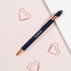 K&ouml;nnte beinhalten: Ein marineblauer Stift mit ros&eacute;goldenen Akzenten und dem Namen "Isabelle" aufgedruckt. Umgeben von drei herzf&ouml;rmigen B&uuml;roklammern in Ros&eacute;gold auf einem hellrosa Hintergrund.