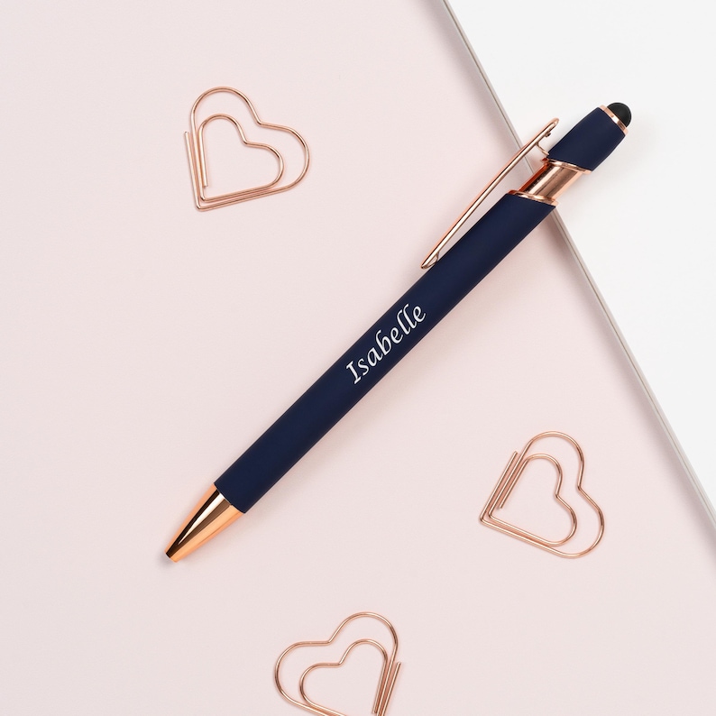 K&ouml;nnte beinhalten: Ein marineblauer Stift mit ros&eacute;goldenen Akzenten und dem Namen "Isabelle" aufgedruckt. Umgeben von drei herzf&ouml;rmigen B&uuml;roklammern in Ros&eacute;gold auf einem hellrosa Hintergrund.