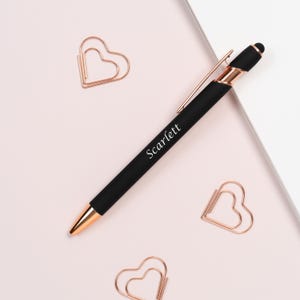 K&ouml;nnte beinhalten: Ein schwarzer Stift mit ros&eacute;goldenen Akzenten und dem Namen "Scarlett" auf dem Schaft. Der Stift liegt auf einer hellrosa Oberfl&auml;che, zusammen mit drei herzf&ouml;rmigen B&uuml;roklammern in Ros&eacute;gold.