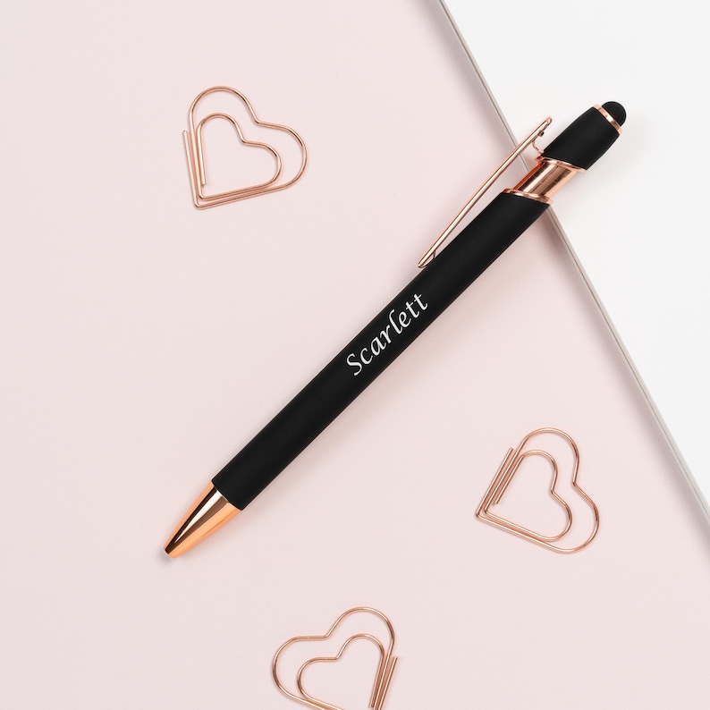 K&ouml;nnte beinhalten: Ein schwarzer Stift mit ros&eacute;goldenen Akzenten und dem Namen "Scarlett" auf dem Schaft. Der Stift liegt auf einer hellrosa Oberfl&auml;che, zusammen mit drei herzf&ouml;rmigen B&uuml;roklammern in Ros&eacute;gold.