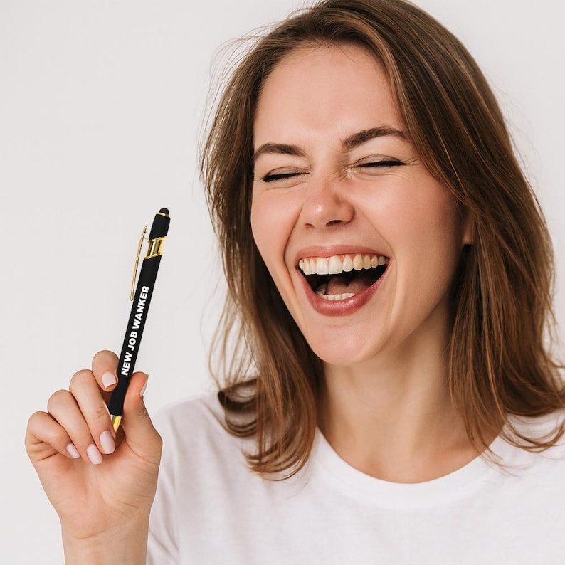 May include: Black and gold pen with the text "NEW JOB WANKER" printed on the barrel. The pen has a stylus tip and gold accents. The pen is held by a person wearing a white t-shirt.