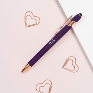 K&ouml;nnte beinhalten: Ein lila Stift mit ros&eacute;goldenen Akzenten und dem Namen "Mila" aufgedruckt. Der Stift liegt zusammen mit drei herzf&ouml;rmigen B&uuml;roklammern auf einer hellrosa Oberfl&auml;che.