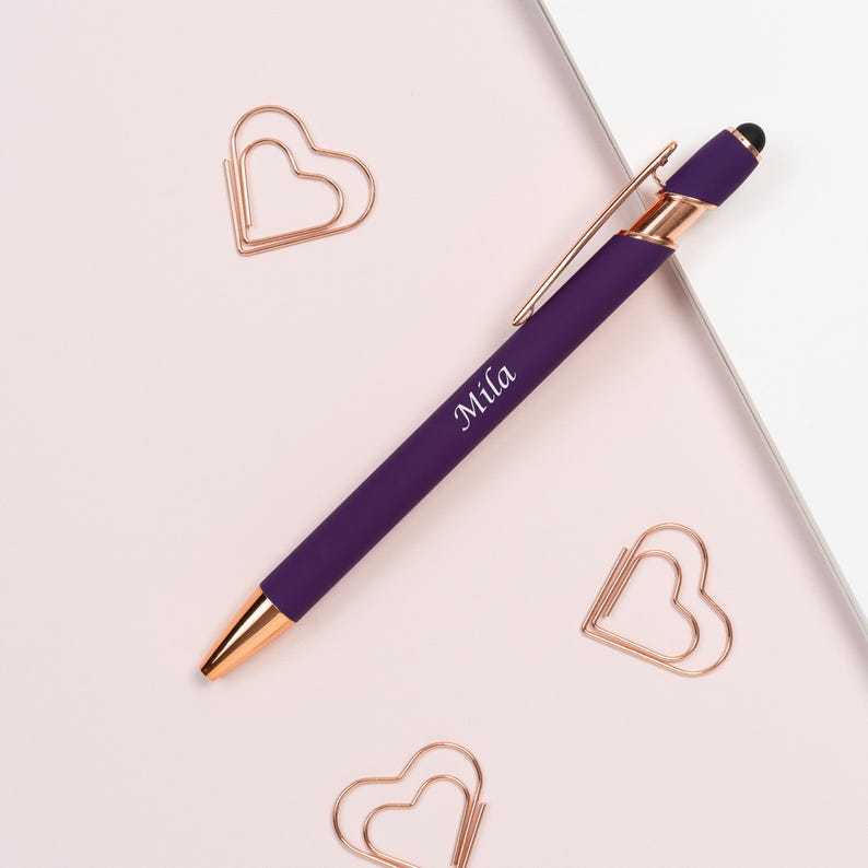K&ouml;nnte beinhalten: Ein lila Stift mit ros&eacute;goldenen Akzenten und dem Namen "Mila" aufgedruckt. Der Stift liegt zusammen mit drei herzf&ouml;rmigen B&uuml;roklammern auf einer hellrosa Oberfl&auml;che.