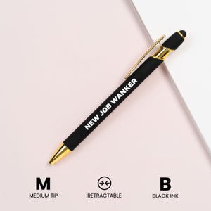 May include: A black retractable pen with gold accents and the text "NEW JOB WANKER" in white. The pen has a medium tip and uses black ink. The pen is on a light pink and white background.