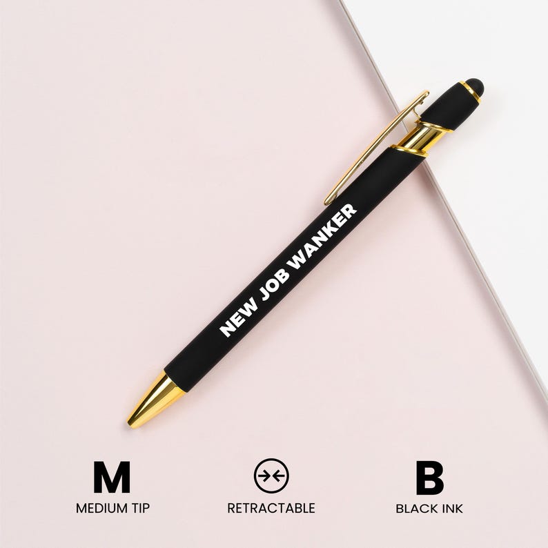 May include: A black retractable pen with gold accents and the text "NEW JOB WANKER" in white. The pen has a medium tip and uses black ink. The pen is on a light pink and white background.