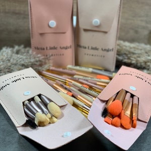Op de afbeelding: Vier rechthoekige cosmetica-etuis in roze en beige tinten, elk voorzien van een witte drukknoopsluiting en de tekst Meta Little Angel Cosmetics. Twee etuis zijn gesloten en twee zijn geopend, met make-up kwasten aan de binnenkant zichtbaar.