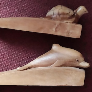 Könnte beinhalten: Zwei Türstopper aus Holz, einer mit einer geschnitzten Schildkröte und der andere mit einem Delfin. Beide sind hellbraun und keilförmig, um Türen offen zu halten. Der Schildkröten-Türstopper hat einen detaillierten Panzer.