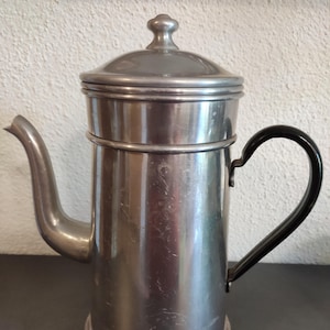 May include: A vintage silver-toned coffee pot with a lid and a black handle. The pot has a spout and a cylindrical body. The handle is curved. The coffee pot is made of metal and has a classic design.