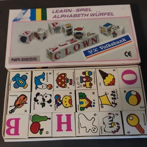 Peut inclure: Un coffret de blocs d'alphabet blancs avec des illustrations colorées et le texte "LEARN-SPIEL ALPHABETH WÜRFEL". L'ensemble comprend des cubes lettrés et des blocs d'images, avec des images comme un clown, une maison et un zèbre. La boîte porte également le texte "Volksbank".