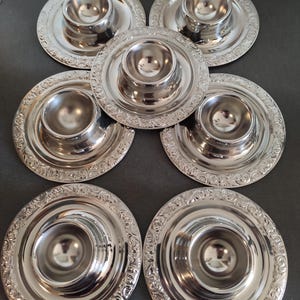 Könnte beinhalten: Sechs silberfarbene Eierbecher mit dekorativen Rändern. Jeder Becher hat eine zentrale Schale für das Ei und eine umlaufende Platte. Das Design ist bei allen sechs Teilen einheitlich.