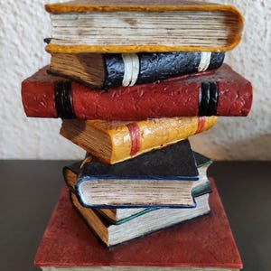 Può includere: Una pila di libri decorativi in vari colori. I libri sono rossi, neri, gialli e verdi, con pagine invecchiate e copertine strutturate. La pila è disposta in una torre leggermente inclinata.