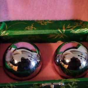 Può includere: Due sfere di metallo argentato in una scatola verde e rosa. Le sfere sono lisce e lucide, e vengono utilizzate per l'esercizio e il rilassamento.