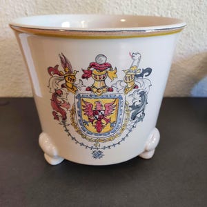 Puede incluir: Jardinera de cerámica color crema con un diseño detallado de escudo de armas. La jardinera tiene una forma ligeramente acampanada, un borde amarillo y tres pequeños pies. El escudo presenta un escudo con un águila roja y otros elementos heráldicos.