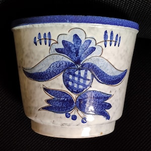 Könnte beinhalten: Ein Keramikübertopf mit gesprenkelter cremeweißer Glasur und einem einfarbig blauen Rand. Die Außenseite zeigt ein handgemaltes blaues Blumenmotiv mit einer ananasartigen Mittelform, symmetrischen Blättern und kleinen Beeren, umrandet von feinen braunen Linien.