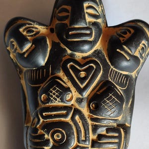 Peut inclure: Une figurine sculptée noire et beige présentant un visage central avec une forme de cœur en dessous, flanqué de deux visages plus petits sur les épaules. La surface est ornée de motifs géométriques, incluant des hachures, des cercles et des lignes.