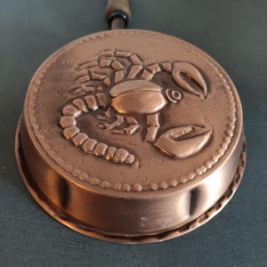 Op de afbeelding: Een koperen pan met een houten handvat en een reliëf van een schorpioen. De pan heeft een gegolfde rand.