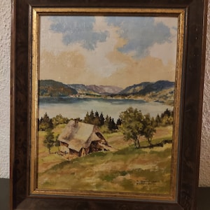 May include: A framed oil painting depicting a landscape scene with a small wooden house in the foreground, a lake in the middle ground, and hills in the background. The sky is a light blue with white clouds. The painting is signed "W. Braun" in the lower right corner.