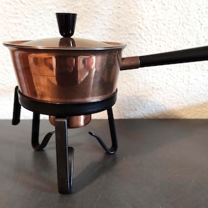 Op de afbeelding: Een koperen fonduepan met een gepolijste afwerking, voorzien van een zwarte dekselknop en een lange zwarte steel. De pan rust op een zwarte metalen standaard met drie gebogen poten, ontworpen om een warmtebron onder de koperen pan te plaatsen.