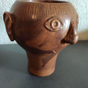 Könnte beinhalten: Eine braune Keramikvase in Form eines menschlichen Gesichts. Die Vase hat einen abgerundeten Kopf mit strukturierter Oberfläche, eine markante Nase, ein Auge und ein Ohr. Der Sockel ist ein ausgestellter Sockel.