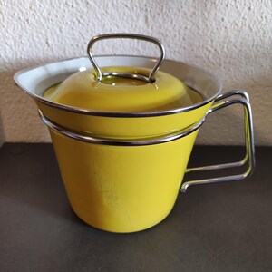 May include: A yellow enamelware pot with a lid and a silver handle. The pot has a silver handle and a white interior. The pot is sitting on a dark surface.