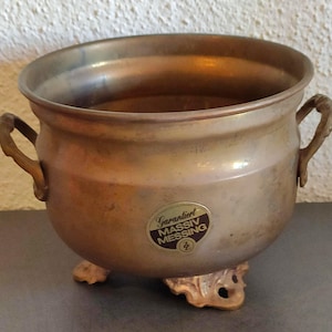 Può includere: Un calderone vintage in ottone con due manici e tre piedi ornati. Il calderone ha un corpo arrotondato e un'ampia apertura. Un adesivo sulla parte anteriore recita "Garantirt Massiv Messing".