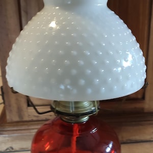 Puede incluir: Una lámpara de aceite de vidrio rojo con una pantalla de vidrio blanco texturizada. La pantalla tiene un borde con cuentas.