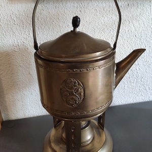 Könnte beinhalten: Eine antike Messing-Teekanne mit einem verzierten Design und einem Holzgriff, die auf einem passenden Messingständer steht.