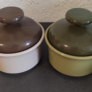 Peut inclure: Deux récipients en céramique avec couvercles. L'un est beige clair avec un couvercle et un bouton marron. L'autre est vert clair avec un couvercle et un bouton vert plus foncé. Les deux ont une forme arrondie.