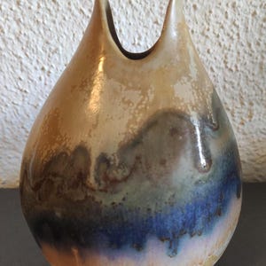 Könnte beinhalten: Eine handgefertigte Keramikvase mit einer einzigartigen, organischen Form. Die Vase hat eine tropfenförmige Form mit zwei nach oben gebogenen Ausgüssen. Die Farbpalette umfasst Brauntöne, Beige, Blau und Grün, wodurch ein Marmoreffekt entsteht. Die Vase ist etwa 20 cm hoch.