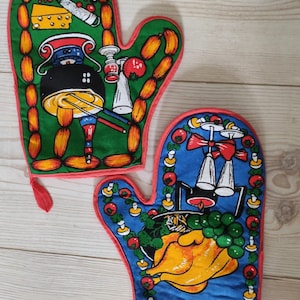 Può includere: Un paio di guanti da forno colorati con un bordo rosso. Un guanto presenta uno sfondo verde con una pentola di cibo, salsicce e formaggio. L'altro guanto ha uno sfondo blu con un pollo arrosto, uva e candele.