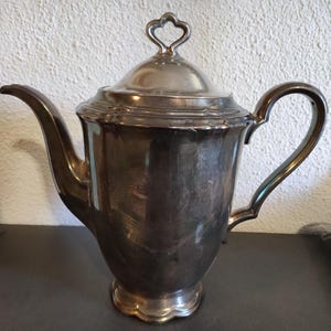 Könnte beinhalten: Eine silberne Teekanne mit einem herzförmigen Knauf auf dem Deckel. Die Teekanne hat ein geriffeltes Design um den oberen Rand und einen runden Korpus.