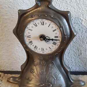 Può includere: Un orologio argentato ornato con numeri romani sul quadrante e un motivo floreale sulla base.