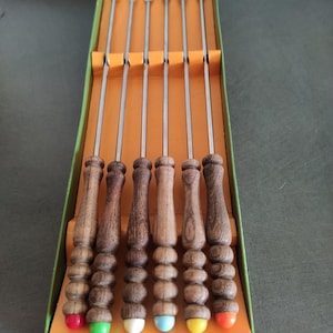 Può includere: Un set di sette forchette da fonduta in una scatola verde e arancione. Le forchette hanno manici in legno con un design bulboso e punte colorate in rosso, verde, bianco, blu, giallo e arancione. La scatola ha del testo in basso.