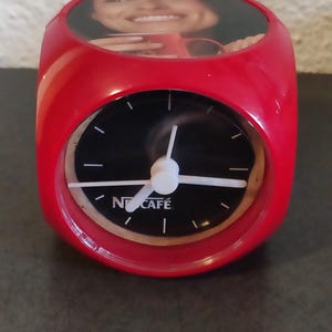 Pode incluir: Um despertador vermelho em forma de cubo com um mostrador preto e ponteiros brancos. O mostrador tem o texto "Nescafé" impresso. A parte superior do relógio apresenta uma imagem de uma pessoa sorrindo segurando uma caneca vermelha.