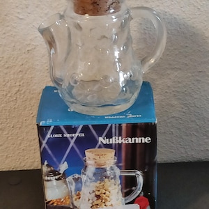 Peut inclure: Une carafe en verre transparent avec un bouchon en liège, posée sur une boîte bleue. La carafe a une poignée et un bec verseur. La boîte porte les inscriptions "GLOBE SHOPPER" et "Nußkanne".
