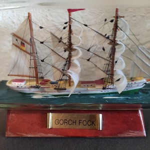 Può includere: Modello dettagliato della nave a vela Gorch Fock all'interno di una bottiglia di vetro. La nave ha vele bianche, alberi marroni e una bandiera rossa. La base è rossa con una targhetta dorata. La nave è ambientata su uno sfondo di onde dipinte.