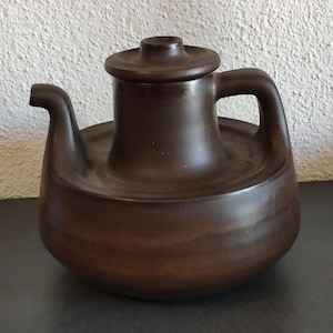 Könnte beinhalten: Eine braune Keramik-Teekanne mit Deckel, Ausguss und Henkel. Die Teekanne hat einen abgerundeten Boden und eine flache Oberseite. Der Griff ist eine gebogene Schlaufe. Die Teekanne ist einfarbig dunkelbraun.