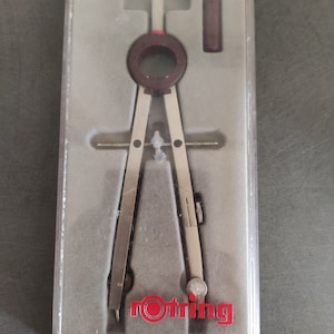 Könnte beinhalten: Ein silberner Zirkel mit einem roten und weißen Knopf und einem roten Rotring-Logo in einem durchsichtigen Kunststoffgehäuse.