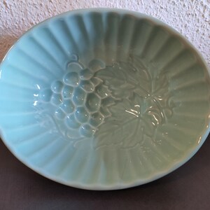 Puede incluir: Un bol de cerámica azul claro con un diseño en relieve de uvas y hojas. El bol tiene un borde acanalado.
