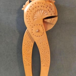 Puede incluir: Un cascanueces de madera marrón claro con intrincados patrones geométricos tallados. El cascanueces tiene una cabeza redondeada con una corona decorativa y una boca articulada. Los mangos son largos y curvos, con tallas detalladas.