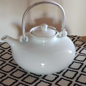 Pode incluir: Uma chaleira de cerâmica branca com uma alça prateada curva e uma tampa a condizer. A chaleira tem um corpo arredondado e um bico. Está sobre uma superfície estampada a preto e branco.