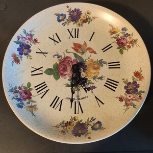 Peut inclure: Une horloge murale ronde avec un cadran crème orné de compositions florales colorées et de chiffres romains noirs. Les aiguilles de l'horloge sont noires, et le texte "EUROPA-QUARTZ" est visible. Le diamètre de l'horloge est d'environ 30 cm.