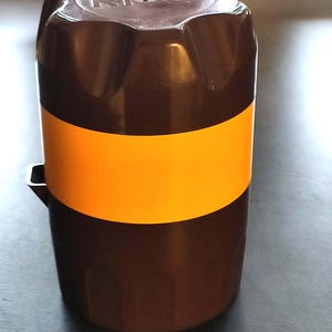 Op de afbeelding: Een bruine en oranje plastic container met het woord "RIO" op de bovenkant. De container heeft een cilindrische vorm met een oranje band in het midden. De bovenkant heeft een geribbeld ontwerp.