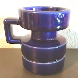 Può includere: Un vaso in ceramica blu cobalto con corpo cilindrico e manico quadrato. Il vaso ha una finitura lucida e un design moderno e geometrico. Il vaso è alto circa 13 cm.